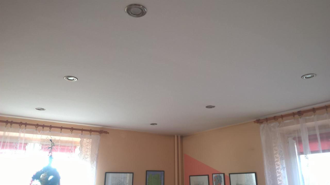 Biały sufit z sześcioma punktowymi lampami halogenowymi, pomarańczowe ściany z obrazami w ramkach i firanki w oknach.