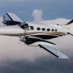 Dwusilnikowy samolot pasażerski w locie, biało-czarna kolorystyka z brązowymi akcentami, rejestracja SP-FNV, widoczne obracające się śmigła na tle chmur.