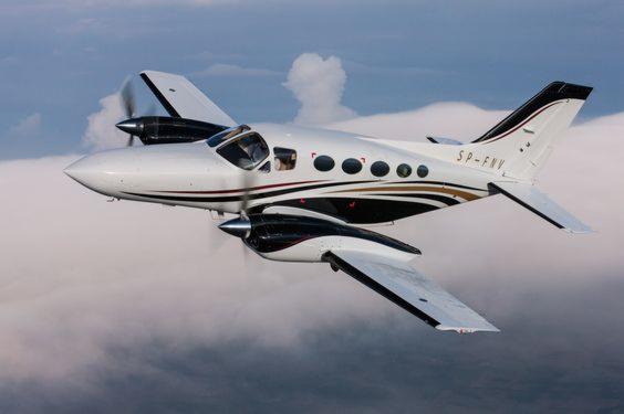 Dwusilnikowy samolot pasażerski w locie, biało-czarna kolorystyka z brązowymi akcentami, rejestracja SP-FNV, widoczne obracające się śmigła na tle chmur.