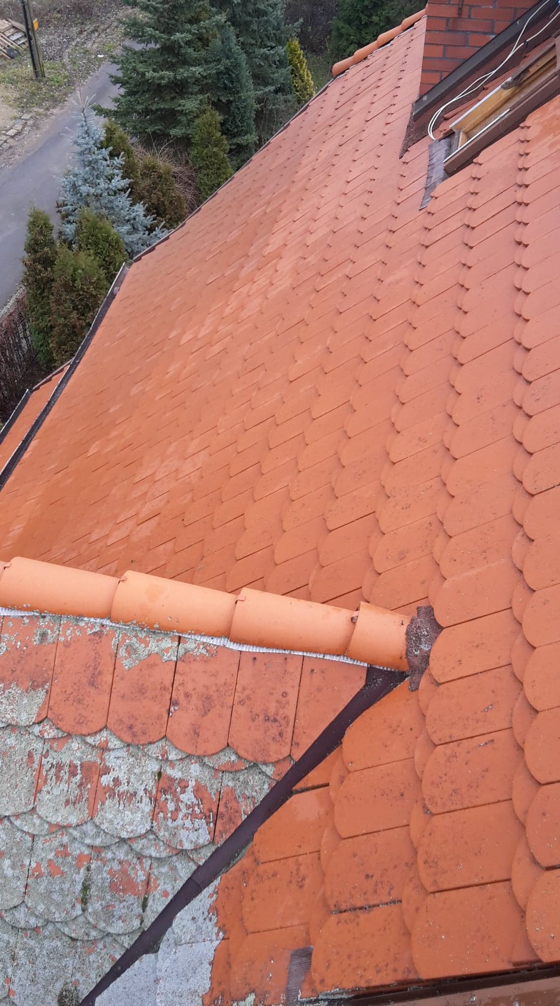 Czerwona dachówka karpiówka na dachu domu, część dachu pokryta starą, szarą dachówką z mchem, w tle zielone drzewa.