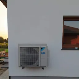 Zewnętrzna jednostka klimatyzacji Daikin zamontowana na białej ścianie budynku, widoczne okno z brązową ramą i fragment tarasu w tle, światło popołudniowe.