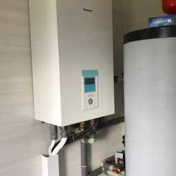 Grupa E-Vento - Biały, wiszący kondensacyjny kocioł gazowy Panasonic z niebieskim wyświetlaczem, zamontowany na kafelkowanej ścianie, z widocznymi rurami instalacyjnymi i zbiornikiem buforowym.