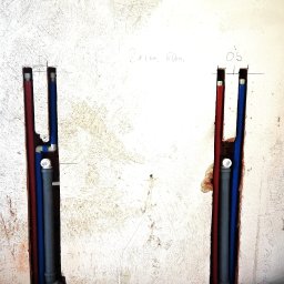 Grupa E-Vento - Wykute w ścianie bruzdy z pionowo poprowadzonymi rurami instalacji wodnej (czerwona i niebieska) oraz kanalizacyjnej (szara), widoczne fragmenty instalacji ogrzewania podłogowego na styropianowej...