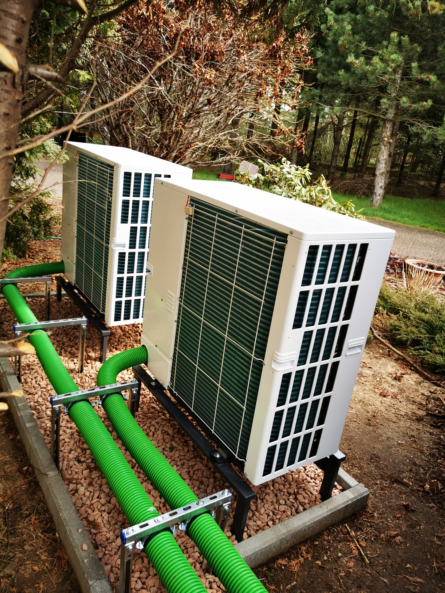 Dwa białe agregaty pompy ciepła zamontowane na zewnątrz budynku, z widocznymi zielonymi rurami doprowadzającymi czynnik chłodniczy, ustawione na żwirowym podłożu w otoczeniu drzew i krzewów.