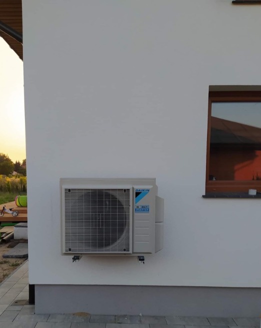 Zewnętrzna jednostka klimatyzacji Daikin zamontowana na białej ścianie budynku, widoczne okno z brązową ramą i fragment tarasu w tle, światło popołudniowe.