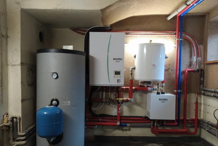 Kotłownia z szarym zbiornikiem buforowym, białym zasobnikiem ciepłej wody i pompą ciepła Ecodan Mitsubishi w surowym pomieszczeniu.