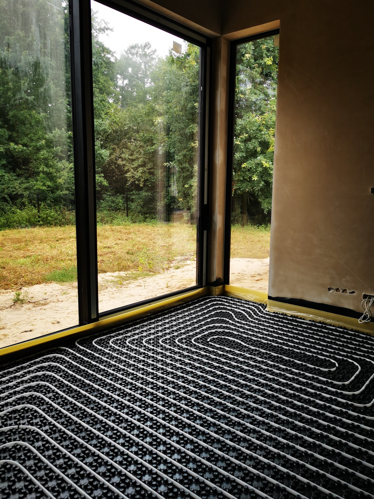 Rozłożona instalacja ogrzewania podłogowego w nowym budynku, widok przez duże okno na zieleń, rury grzewcze wpięte w czarne maty mocujące, zabezpieczone folią krawędzie przy ścianach.