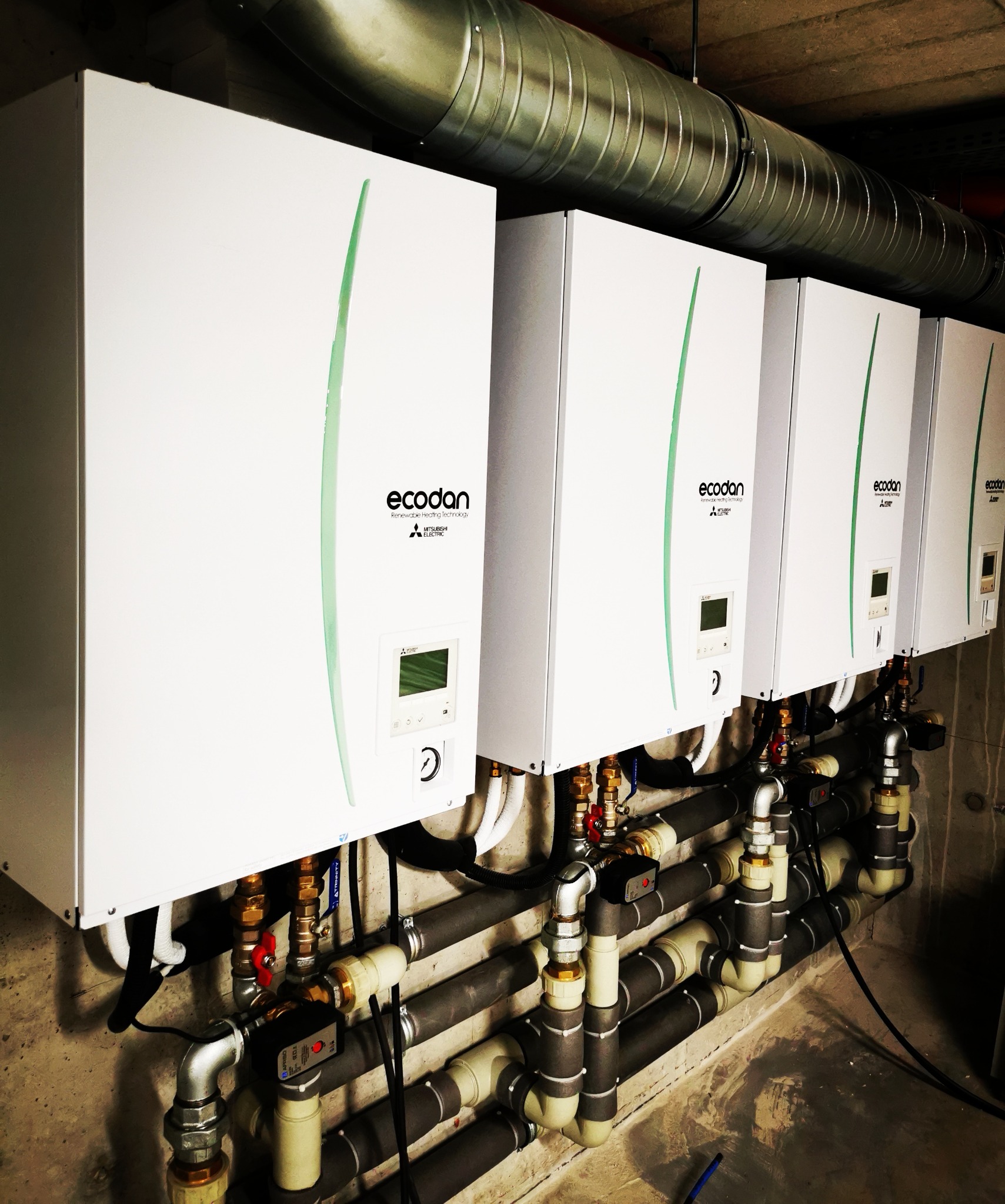 Rząd czterech białych jednostek grzewczych Ecodan Mitsubishi Electric zamontowanych na ścianie, z widocznymi rurami i zaworami, oraz kanałem wentylacyjnym pod sufitem.
