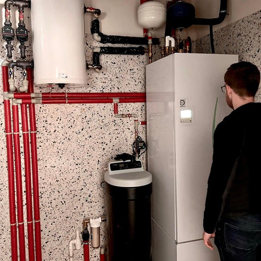 Instalacja kotła grzewczego z podłączonymi rurami, zmiękczaczem wody i zbiornikiem wyrównawczym w pomieszczeniu z mozaikową ścianą, widziana z perspektywy osoby stojącej obok.