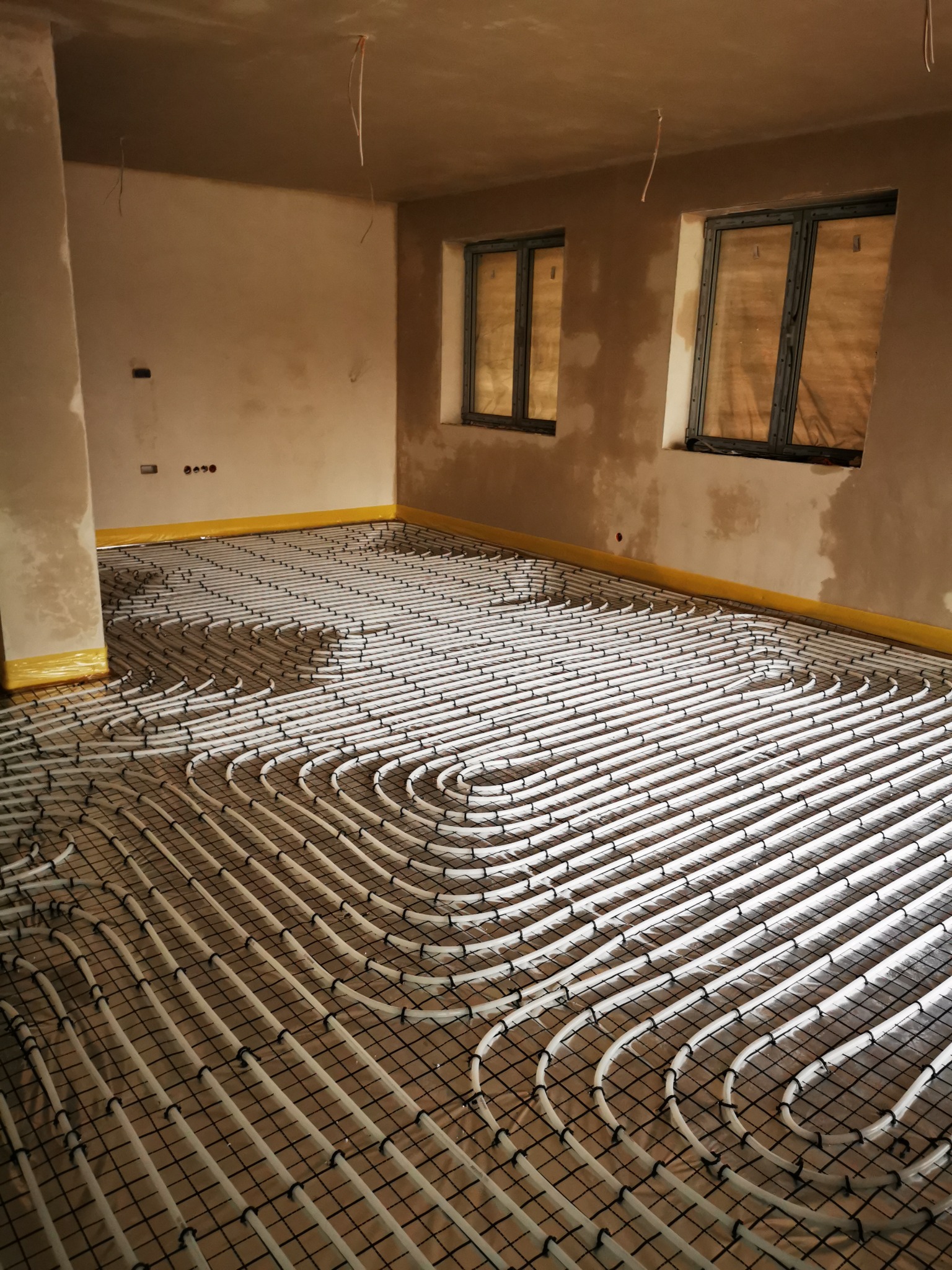 Rozłożona instalacja ogrzewania podłogowego na siatce w surowym pomieszczeniu z dwoma oknami przed wylewką.