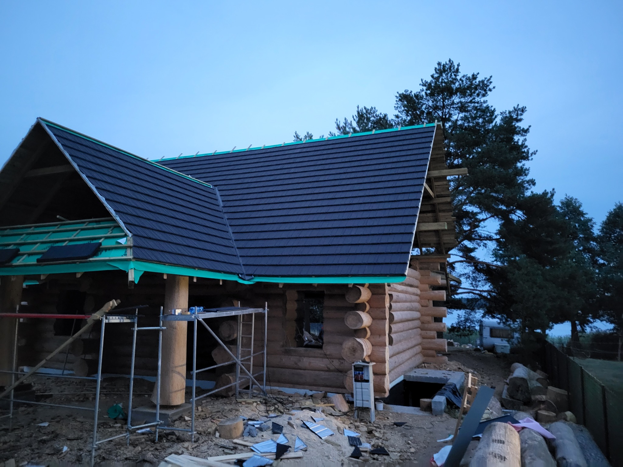 Budowa domu z bali w trakcie krycia dachu blachodachówką w kolorze grafitowym, widoczne rusztowania i przygotowane elementy więźby dachowej.