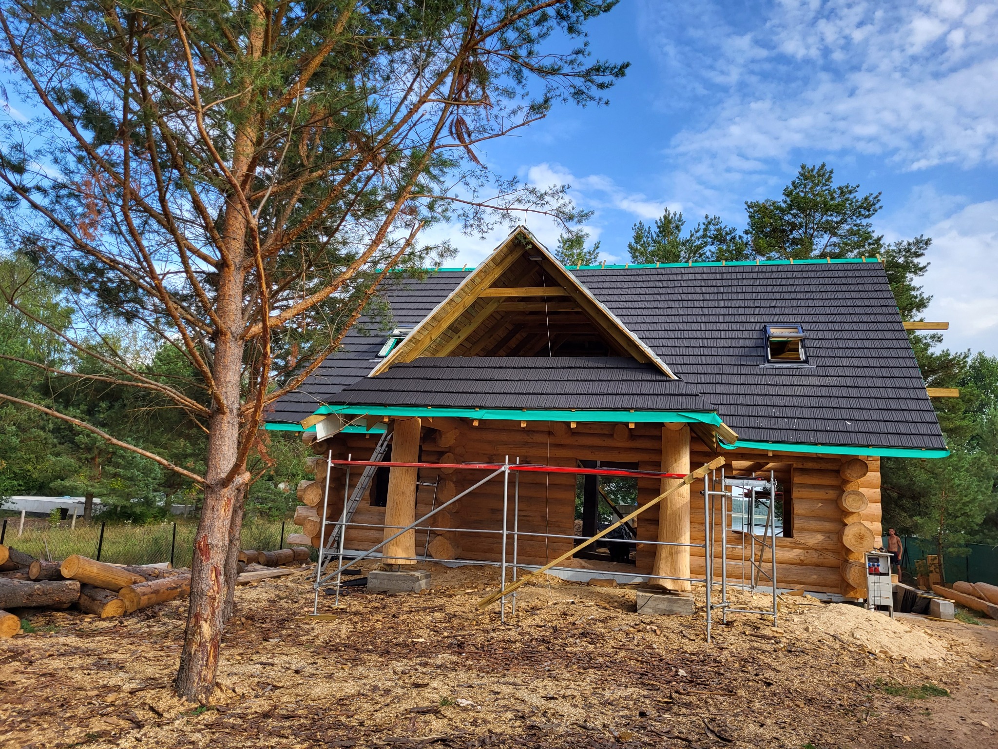 Budowa domu z bali z częściowo ukończonym dachem z czarnej dachówki, widoczne rusztowania, stos drewna i wysokie drzewo na pierwszym planie.