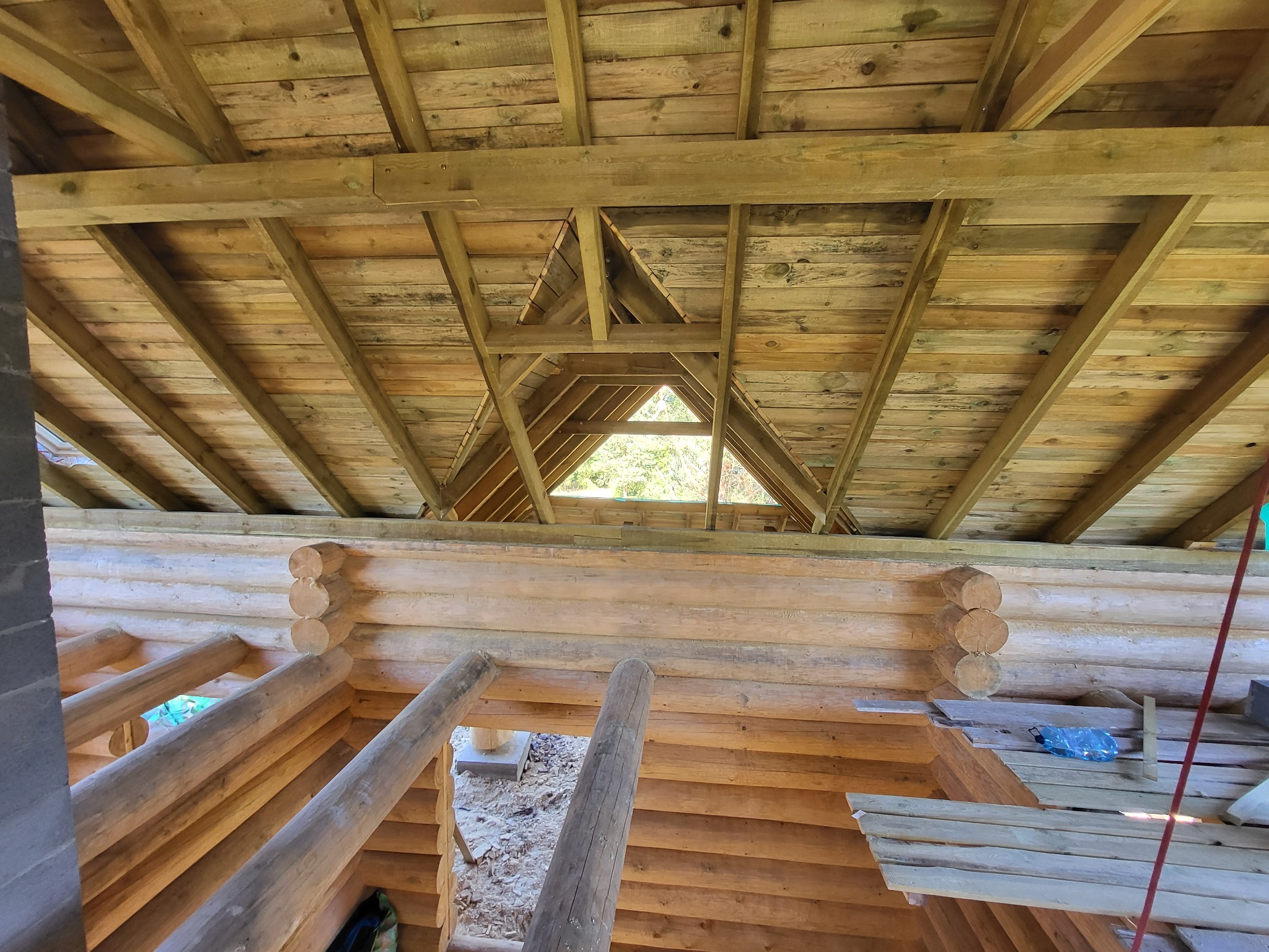 Wnętrze drewnianego domu w trakcie budowy z widoczną więźbą dachową i ścianami z bali, z trójkątnym otworem w dachu przepuszczającym światło.