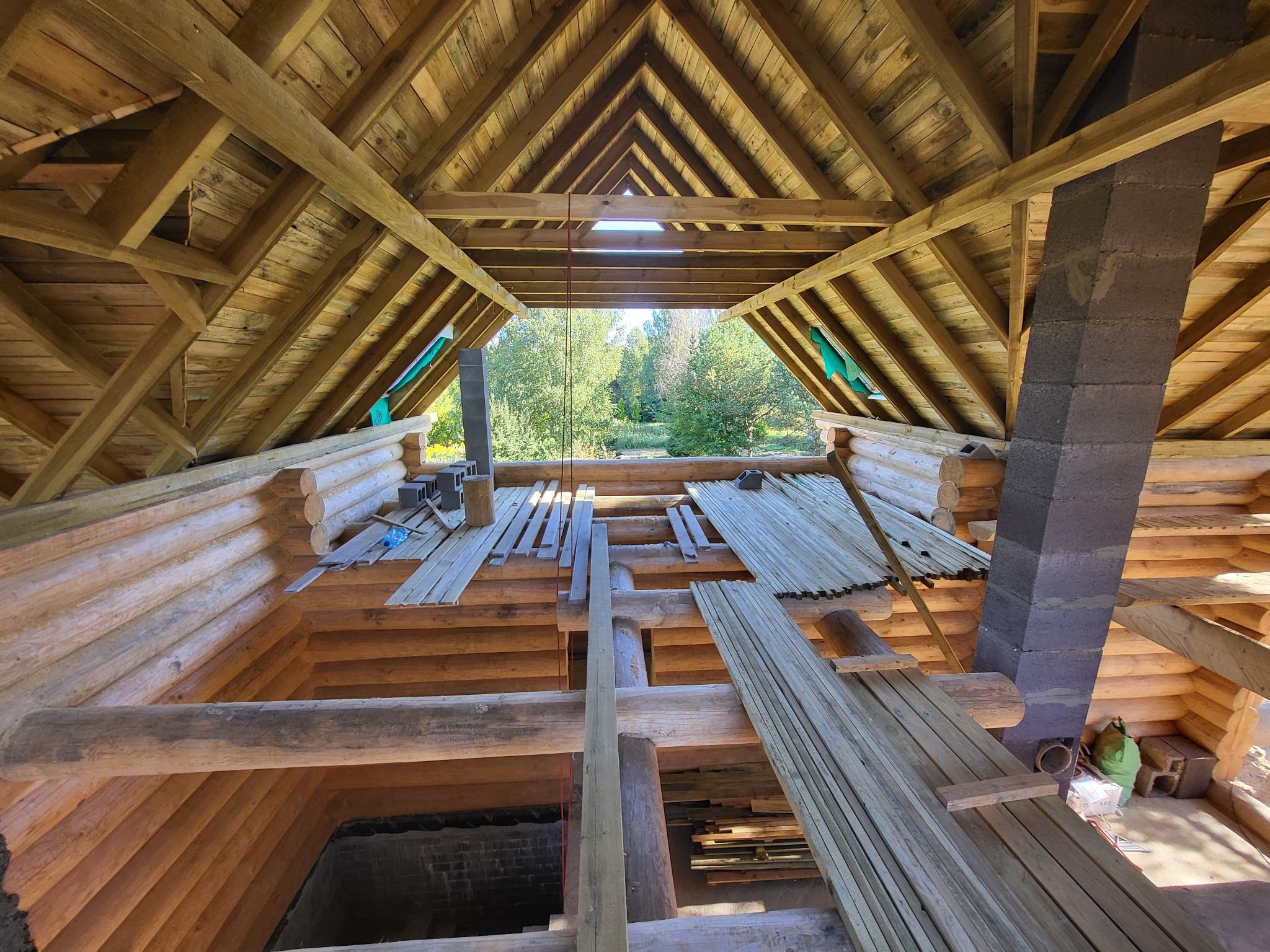 Wnętrze drewnianego domu z widoczną konstrukcją dachu i kominem w trakcie budowy, widok na zewnątrz przez otwór w dachu.
