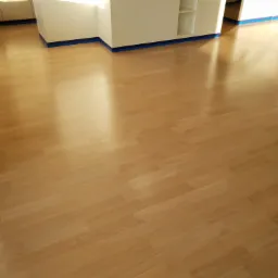 Jasna, nowo położona podłoga z parkietu w pokoju z białymi ścianami i niebieską taśmą malarską wzdłuż krawędzi, odbijająca światło słoneczne.