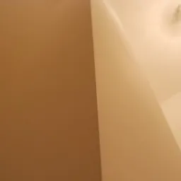 Fragment wnętrza z gładką, beżową ścianą i sufitem, oświetlonym przez żarówkę. Widoczny kąt ściany i sufitu, z delikatnym cieniem.