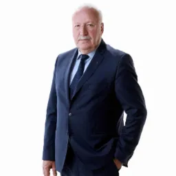 Portret starszego mężczyzny w granatowym garniturze i krawacie, z ręką w kieszeni, na białym tle.