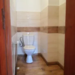 Toaleta w wykończonej łazience, widok z perspektywy wejścia, beżowo-brązowe płytki na ścianach i podłodze imitujące drewno, biała ceramika sanitarna.