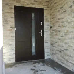 Ciemnobrązowe drzwi wejściowe z przeszkleniem bocznym, otoczone dekoracyjną okładziną ścienną imitującą kamień w odcieniach beżu i szarości. Na posadzce widoczne ślady wilgoci.