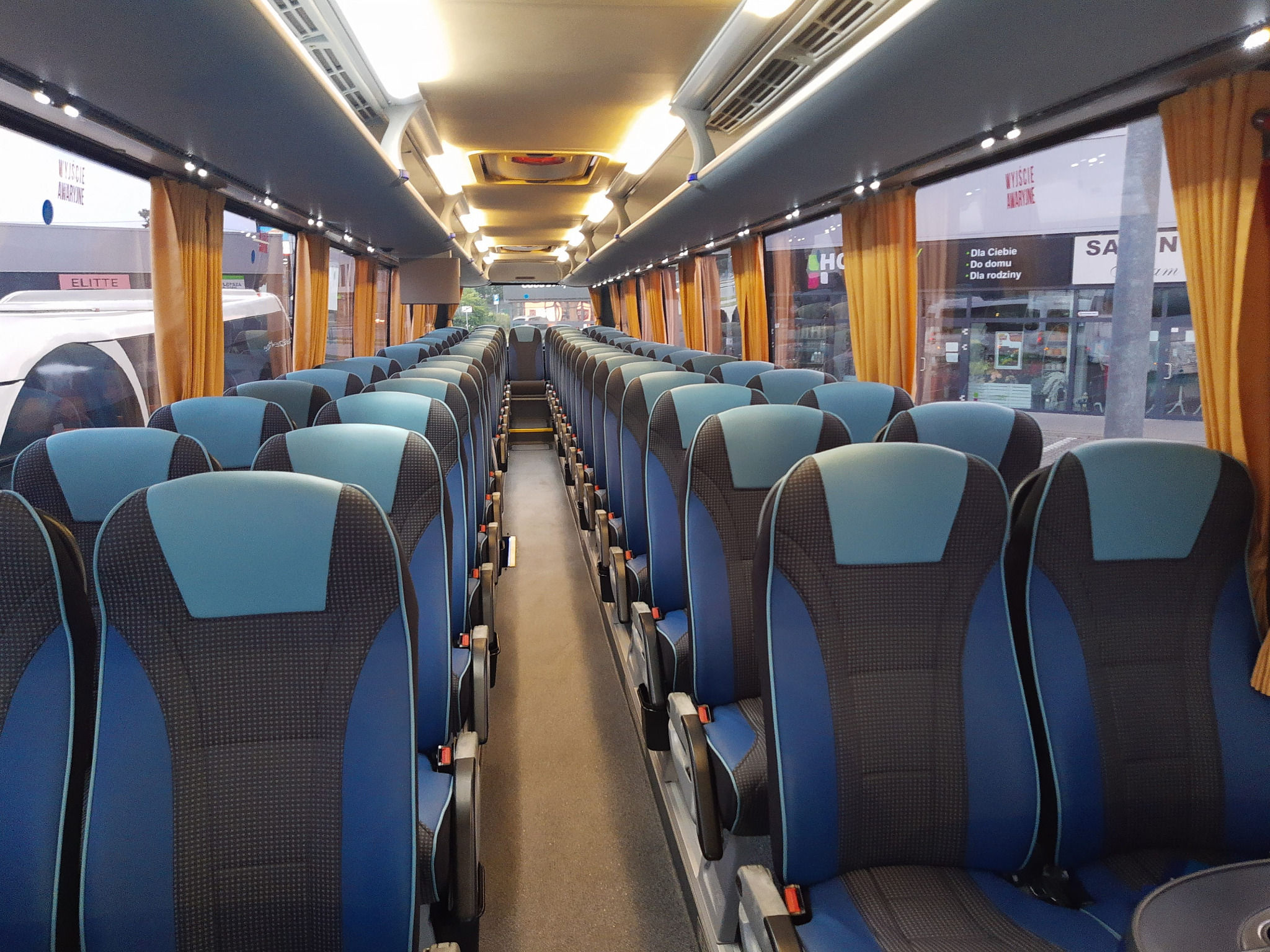 Wnętrze autobusu turystycznego Neoplan Tourliner z rzędami niebiesko-czarnych foteli, widok na przejście w kierunku przodu pojazdu, z widocznymi zasłonami w oknach.