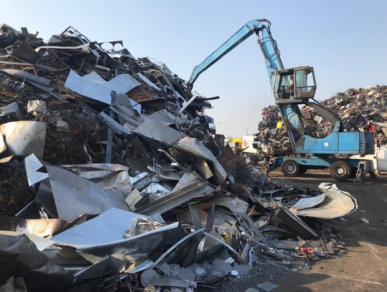 Niebieski dźwig manipuluje stertą złomu metalowego na placu składowym, widoczne fragmenty karoserii samochodowych i blachy.