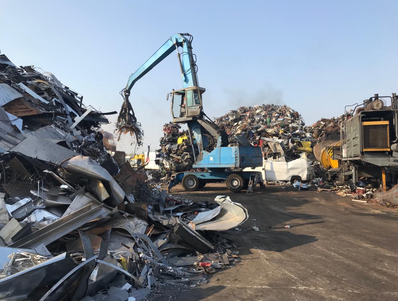 Niebieski dźwig chwytakowy przenosi złom metalowy na składowisku, w tle hałdy złomu i pojazdy.