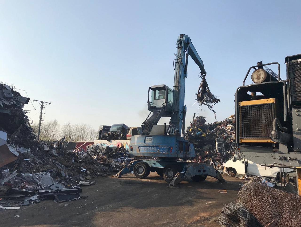 Niebieski dźwig chwytakowy przenosi złom na składowisku, widoczne zgniecione blachy i inne odpady metalowe.
