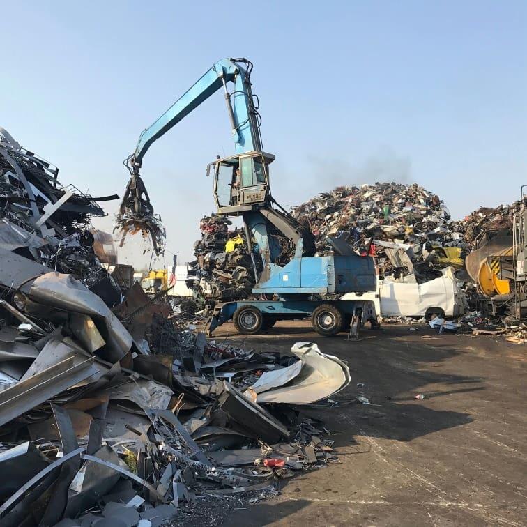Niebieski dźwig chwyta złom metalowy na składowisku, w tle widoczne hałdy odpadów i pojazdy.