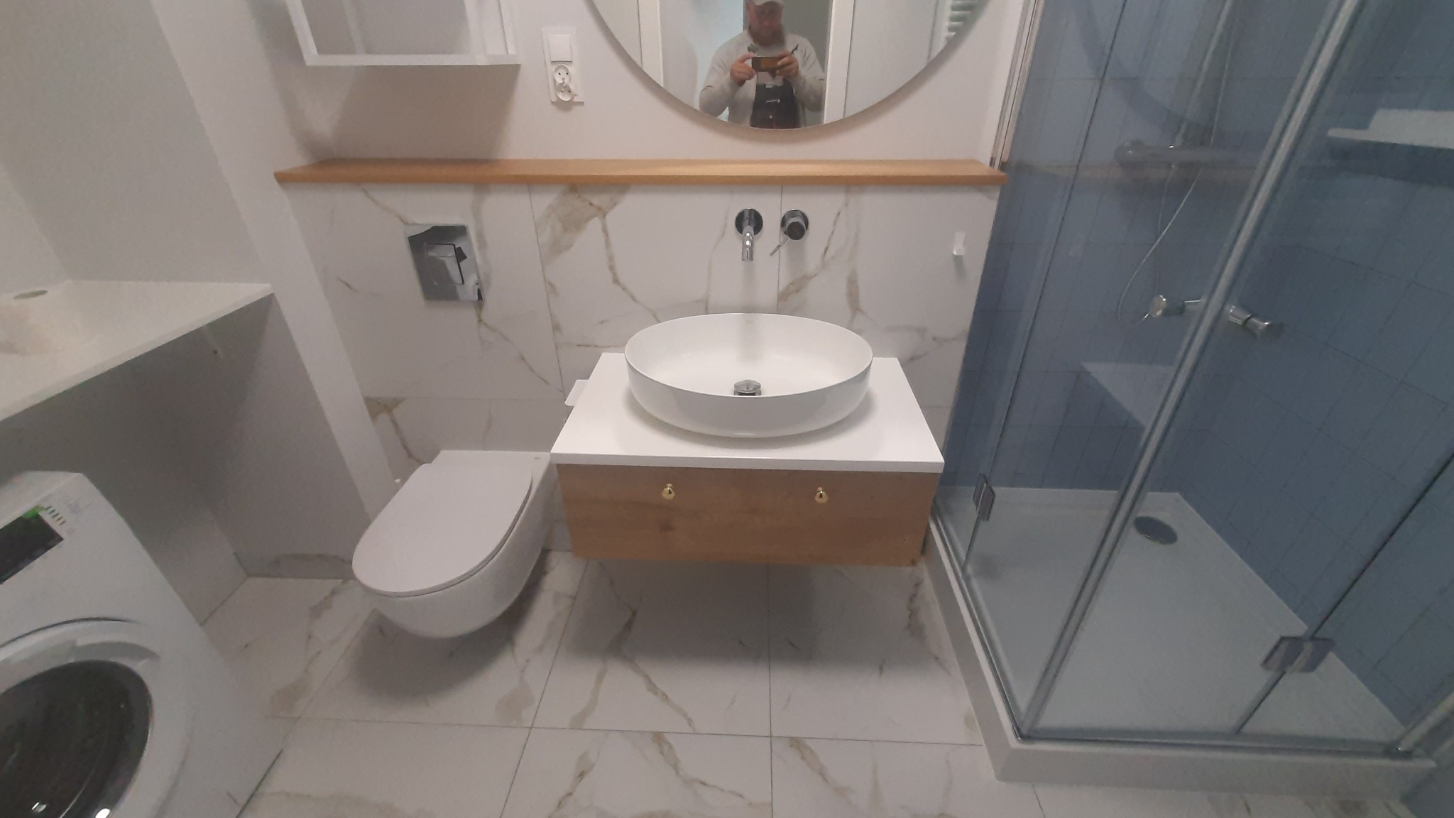 Nowoczesna łazienka z marmurowymi płytkami, okrągłą umywalką nablatową na drewnianej szafce z złotymi uchwytami, kabiną prysznicową ze szklanymi drzwiami i pralką z lewej strony.