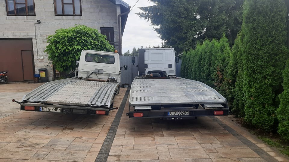 Dwa białe samochody lawety zaparkowane na brukowanym podjeździe przed budynkiem i żywopłotem.