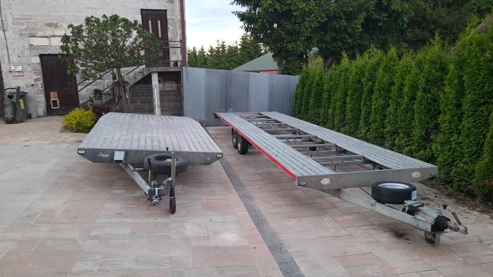 Dwie przyczepy lawety stojące na brukowanym placu w Tarnowie, jedna z pełną platformą, druga szkieletowa, na tle zielonego żywopłotu i budynku gospodarczego.