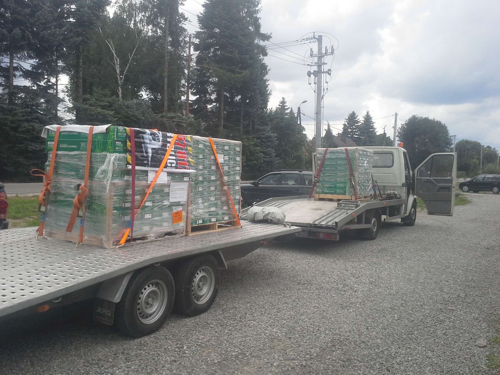 Dwa ładunki palet z materiałami budowlanymi zabezpieczone pasami na platformie przyczepy samochodowej, z otwartymi drzwiami kabiny i widocznym otoczeniem: drzewa, słup elektryczny, samochody...