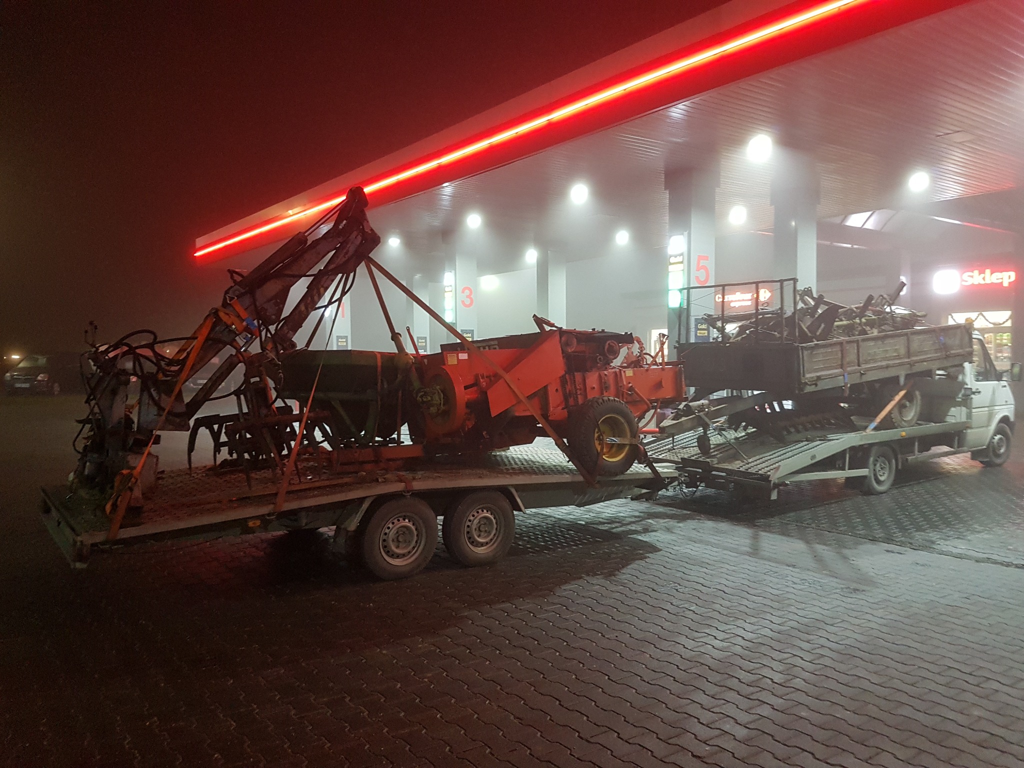 Ciężki sprzęt rolniczy, w tym ciągnik z ładowaczem i przyczepa z elementami, przewożony na platformie lawety w warunkach nocnych na stacji benzynowej, oświetlonej neonowym światłem.