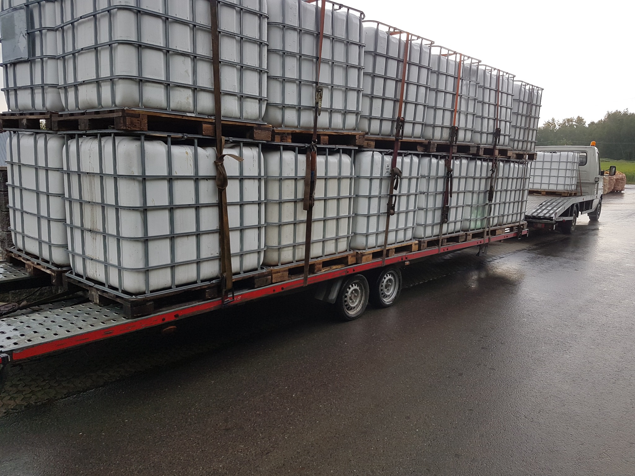 Ciężarówka z przyczepą przewożąca białe kontenery IBC na paletach, zabezpieczone pasami transportowymi, widok z boku na mokrym asfalcie.