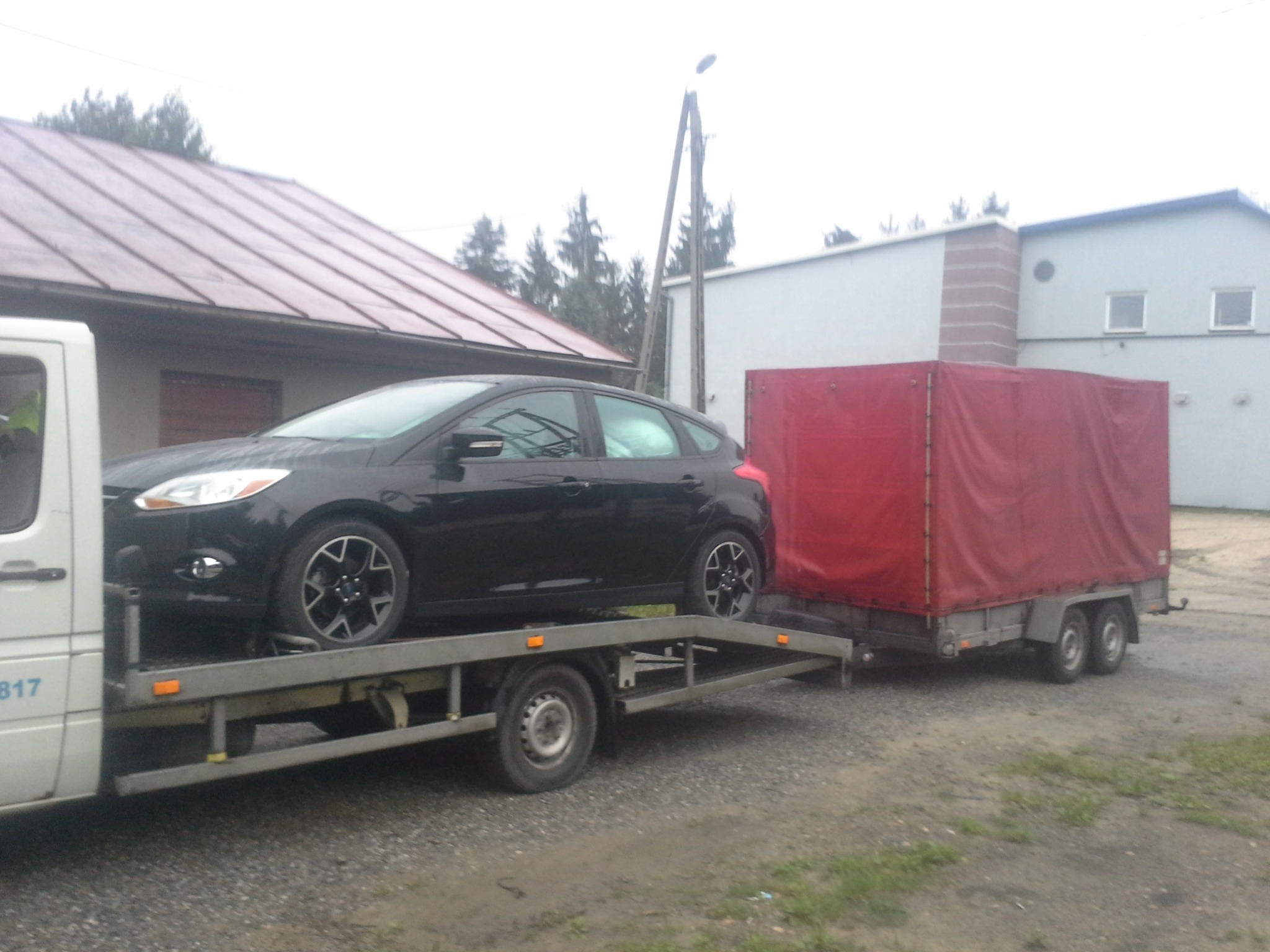Czarny samochód osobowy na platformie transportowej lawety z czerwoną przyczepą w tle zabudowań.