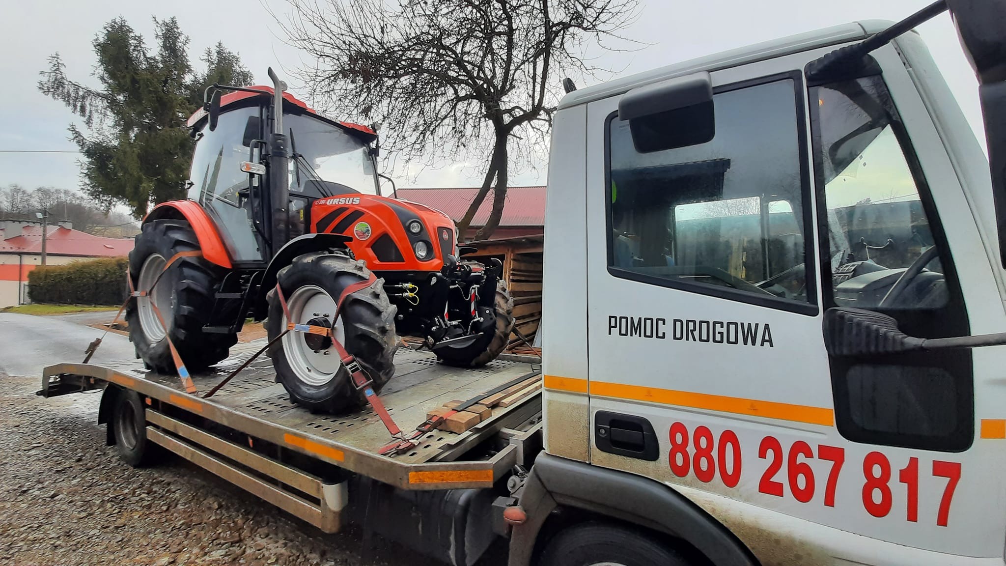 Pomoc Drogowa w Tarnowie transportuje pomarańczowy traktor marki Ursus na lawecie. Na boku lawety widoczny numer telefonu.