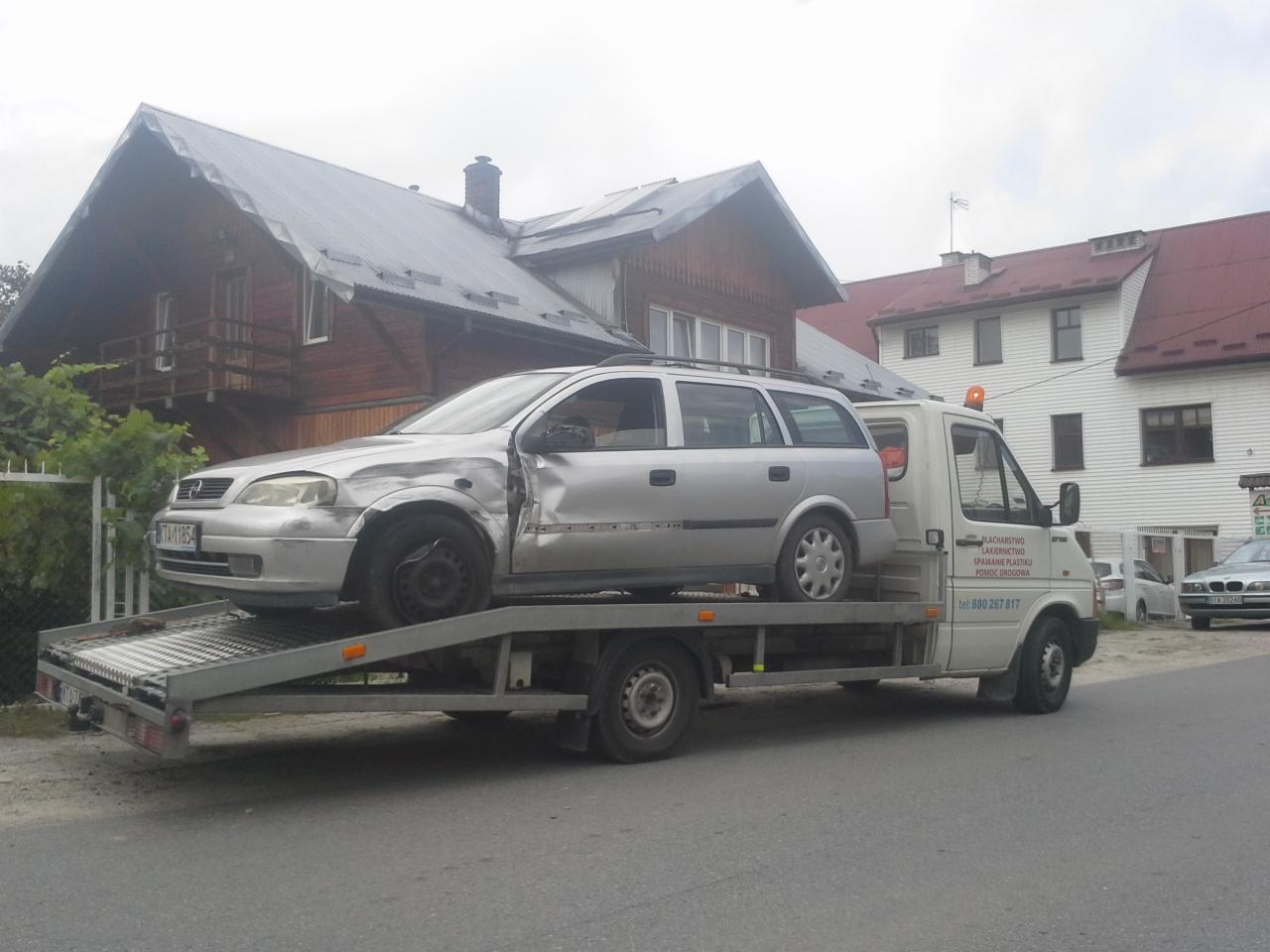 Srebrny samochód osobowy Opel Astra z uszkodzeniami karoserii, bez przedniego koła, załadowany na platformę pojazdu pomocy drogowej na tle zabudowań miejskich.