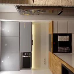 Nowoczesna kuchnia z szarymi, matowymi frontami szafek, drewnianym blatem i wbudowanym piekarnikiem oraz mikrofalówką, oświetlona punktowymi lampami sufitowymi.