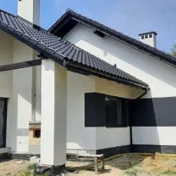 Nowoczesny dom jednorodzinny z białą elewacją i czarnymi akcentami, widoczny fragment ogrodu i elementy wykończeniowe w trakcie realizacji.