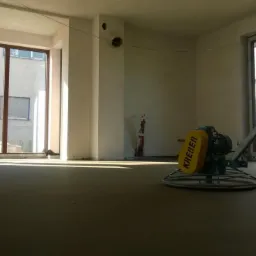 Wnętrze w stanie deweloperskim z maszyną do zacierania posadzek marki Kreber, przygotowane do dalszych prac wykończeniowych, widoczne duże okna i surowe ściany.