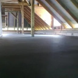 Wylany betonowy podkład podłogowy na poddaszu z drewnianą konstrukcją więźby dachowej i częściowo ocieplonymi ścianami.