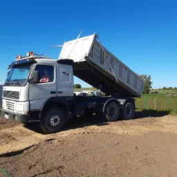 Ciężarówka wywrotka marki Volvo z podniesioną skrzynią ładunkową, stojąca na placu budowy z widoczną ziemią i w tle wiatrak.