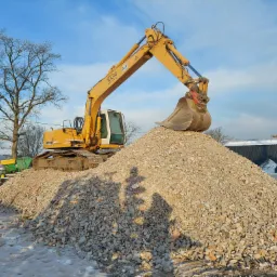 Żółta koparka Liebherr z łyżką na szczycie pryzmy pokruszonego betonu i cegieł, z widocznym śniegiem na ziemi, drzewami bez liści i budynkiem w tle.