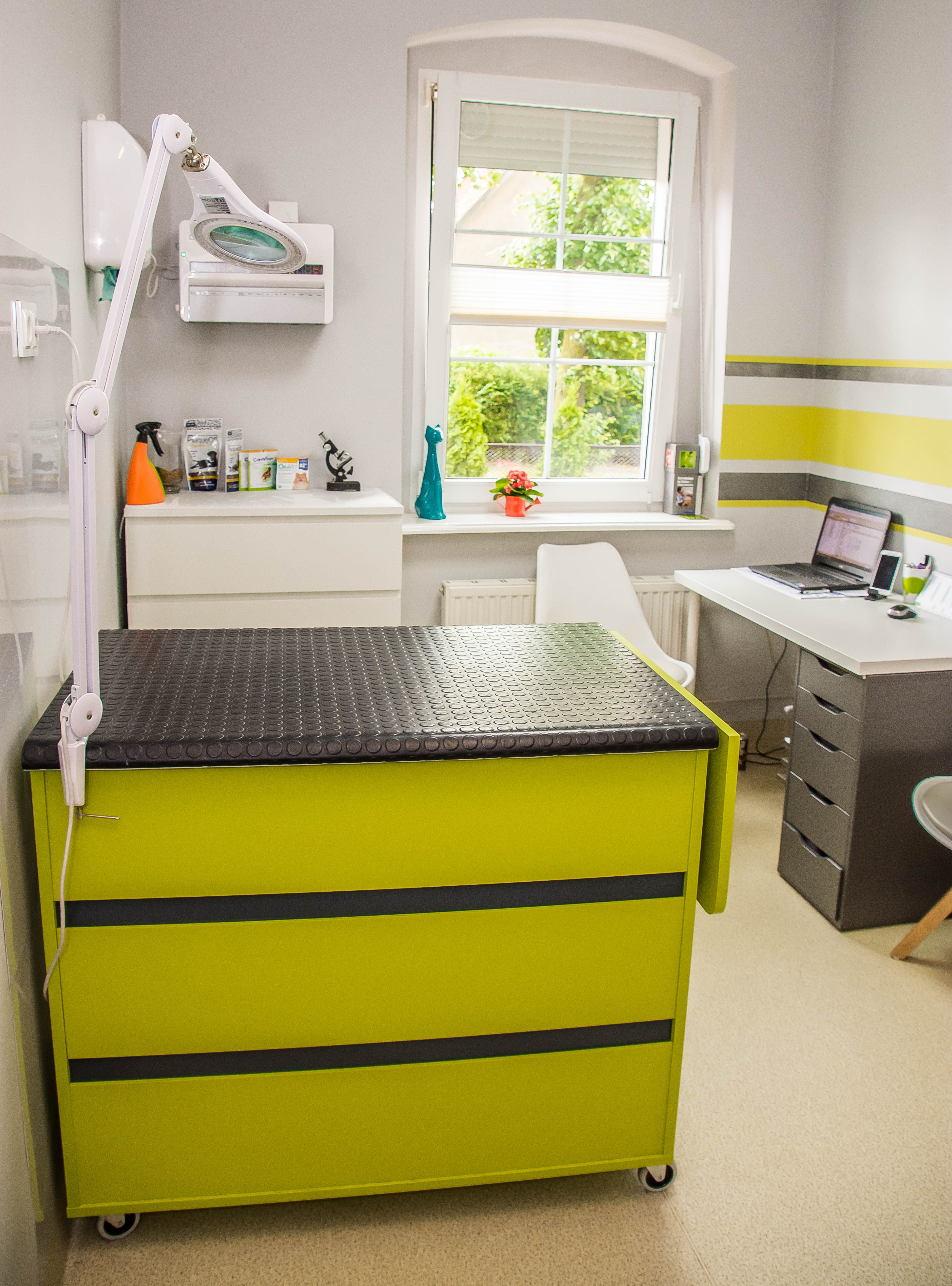 Jasne wnętrze gabinetu z zielono-czarnym stołem zabiegowym na kółkach, lampą powiększającą i biurkiem z laptopem w tle.