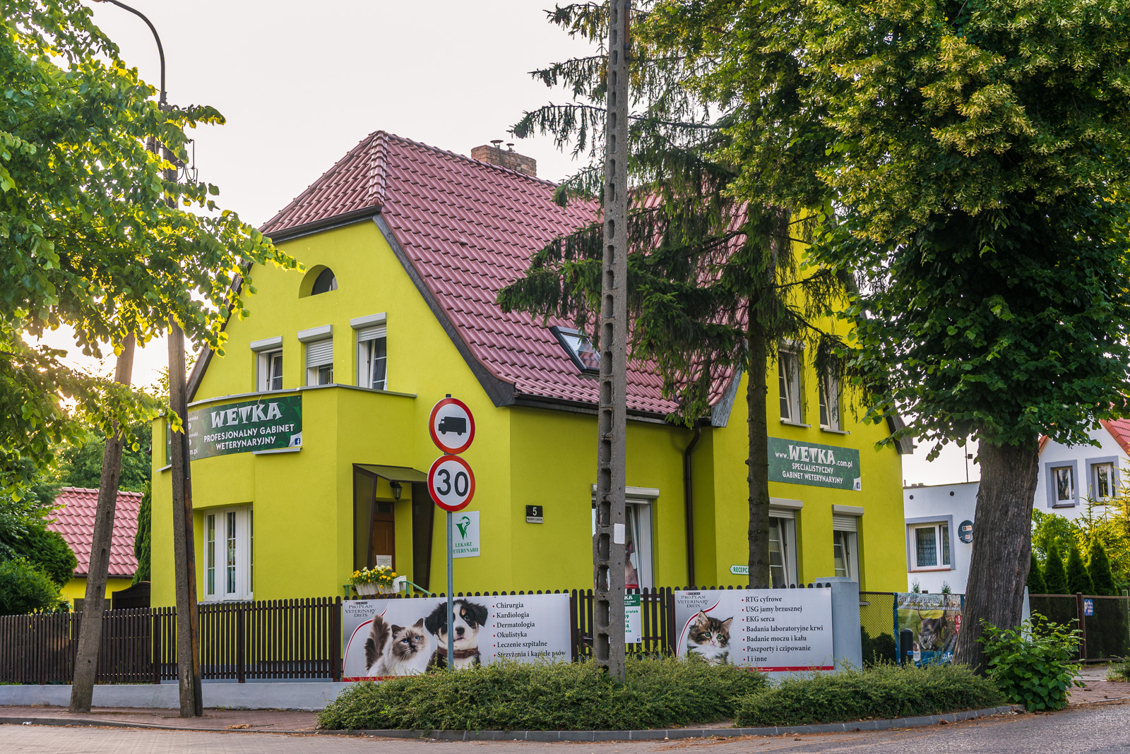 Żółty budynek gabinetu z czerwonym dachem, szyld z nazwą gabinetu oraz banery reklamowe z wizerunkami zwierząt i listą usług, znak ograniczenia prędkości 30 km/h.