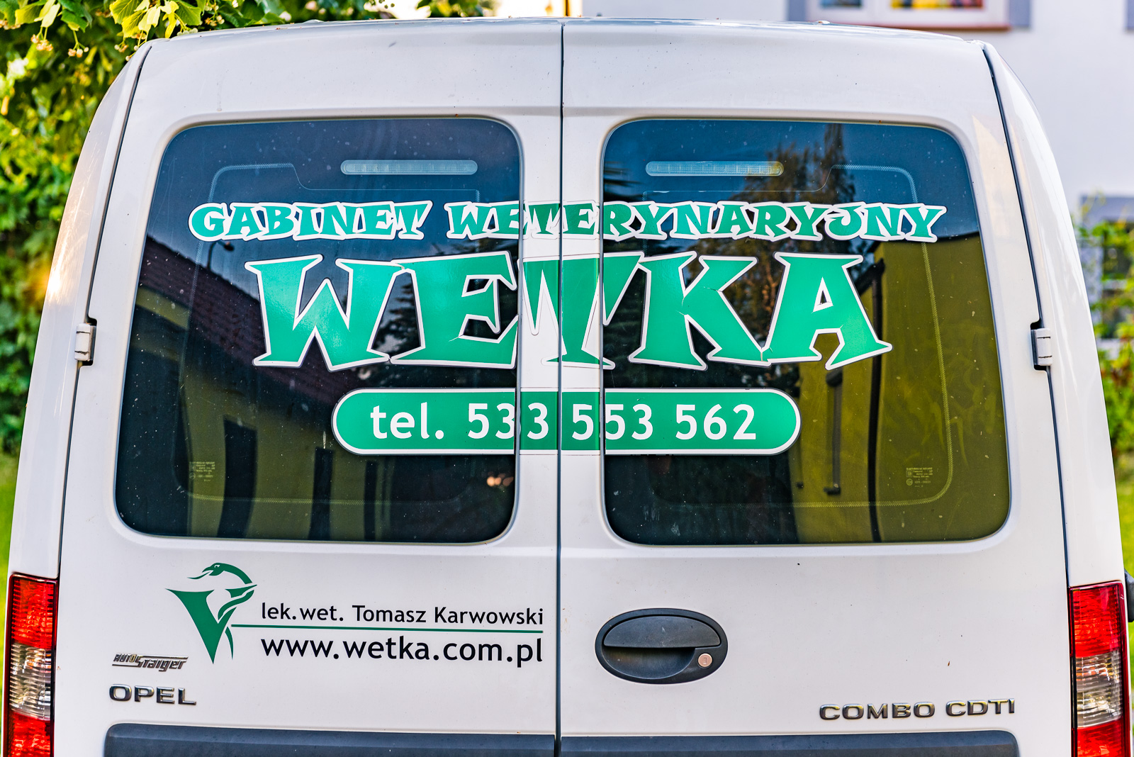 Tylne drzwi białego samochodu dostawczego z naklejkami informującymi o gabinecie weterynaryjnym, nazwa 'Wetka', numer telefonu i adres strony internetowej.