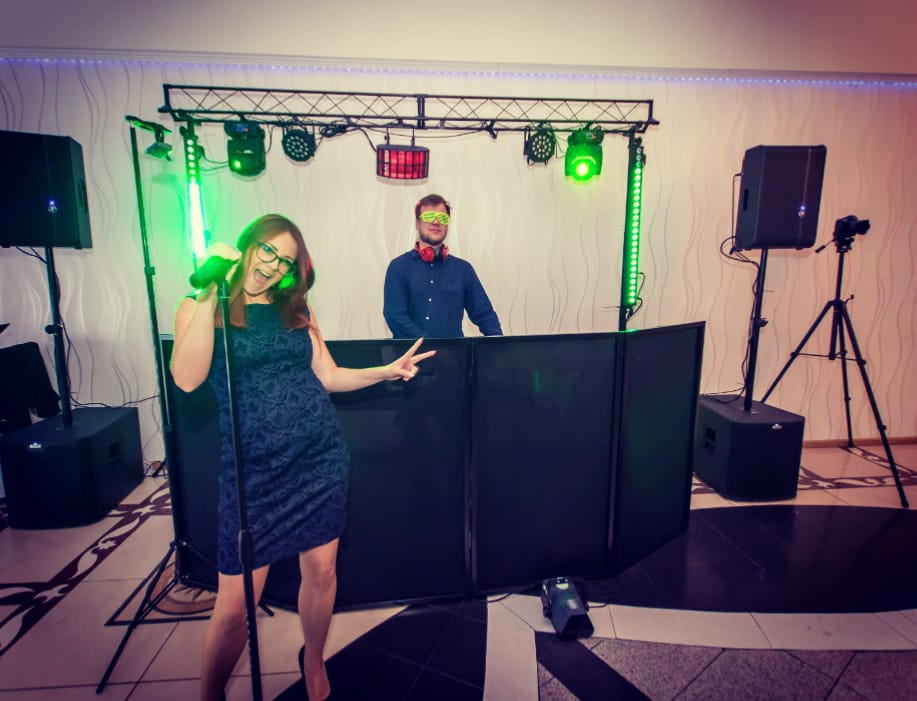 Kobieta w ciemnej sukience śpiewa do mikrofonu, pokazując znak V, za nią DJ w słuchawkach i okularach, oświetlenie sceniczne w tle.