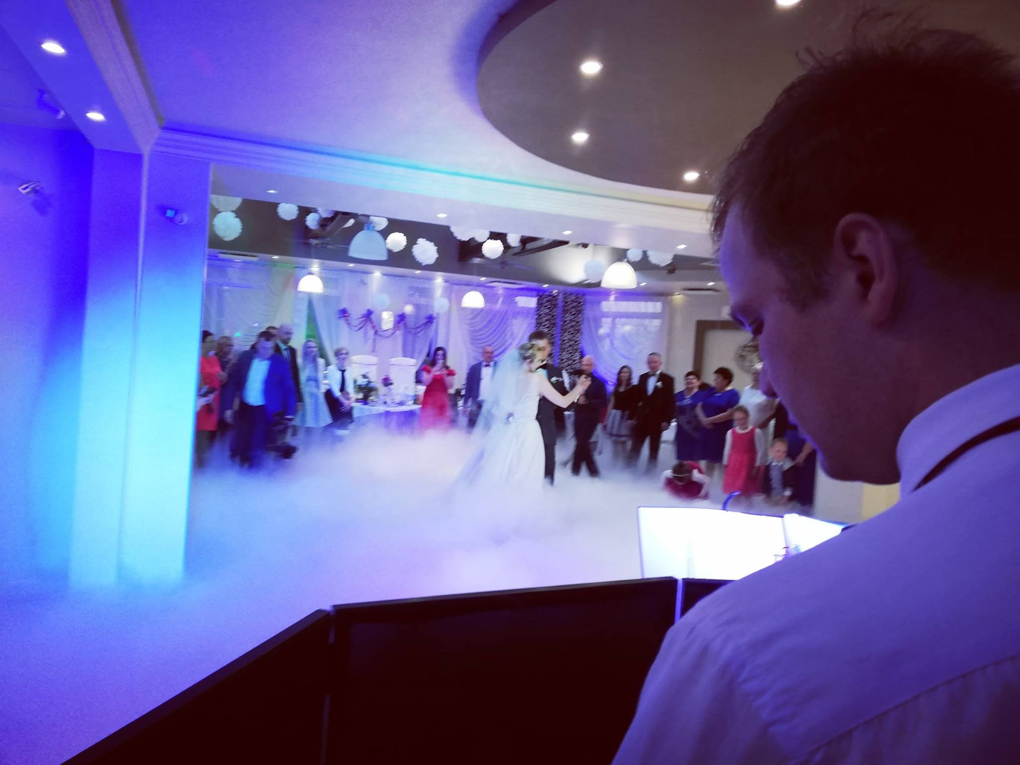 Pierwszy taniec pary młodej w chmurach dymu, widziany zza konsolety DJ-a, w tle goście weselni i dekoracje sali.