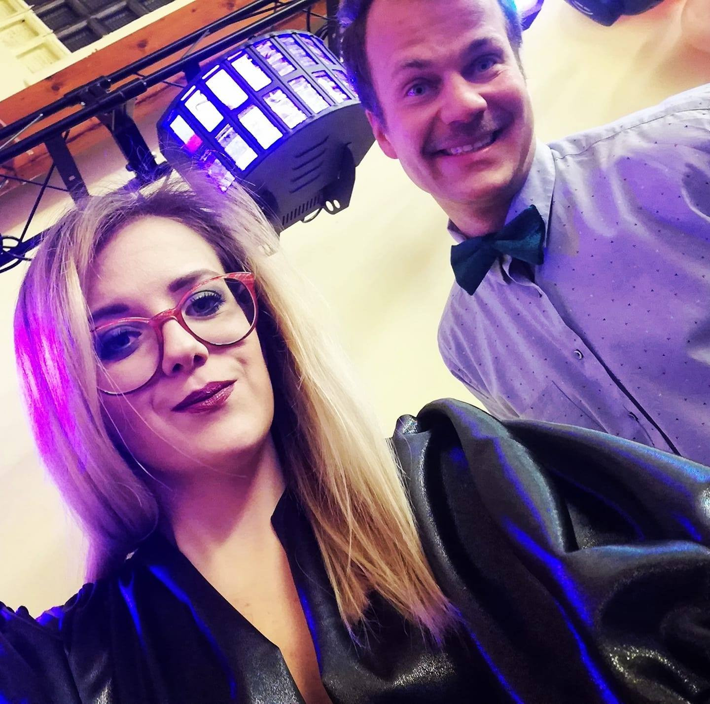 Portret kobiety w okularach i mężczyzny w muszce pod oświetleniem dyskotekowym, oboje uśmiechnięci, z widoczną lampą LED na suficie.