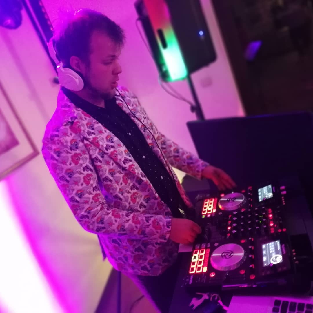 DJ w wzorzystej marynarce i słuchawkach, obsługujący konsoletę DJ-ską oświetloną fioletowym światłem.